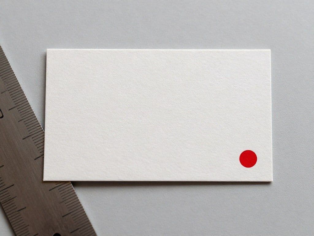 Eine Visitenkarte aus Baumwollpapier mit rotem Punkt, daneben ein Stahllineal.